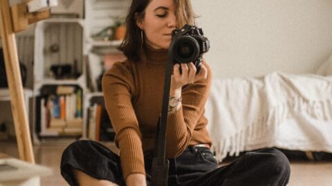 woman sitting on brown wooden floor while holding black DSLR camera in room